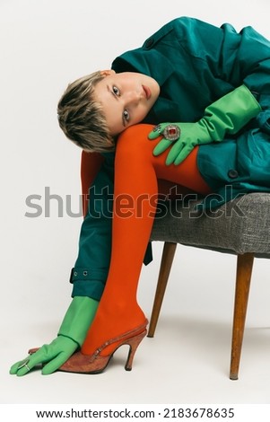 Stylish young girl in bright outfit with green coat, rubber gloves and orange tights sitting on chair isolated over grey background. Concept of retro fashion, art photography, style, beauty
