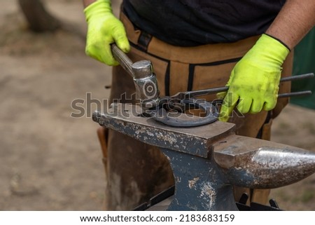 Blacksmith forging a horseshoe with a hammer onto anvil at the foreground. Forging furnace. Farrier fits hot horseshoe for horse. Changing a horseshoe. Blacksmith working with a horse. copy space Royalty-Free Stock Photo #2183683159