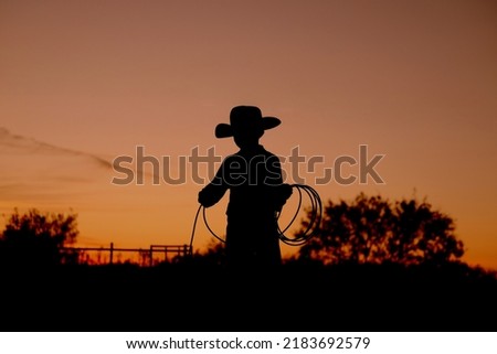 Western industry concept with kid cowboy practicing roping for rodeo at sunset in silhouette on Texas ranch.