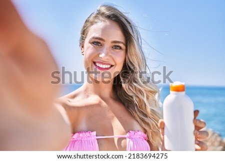 Young blonde girl holding sunscreen lotion make selfie by the camera at the beach.