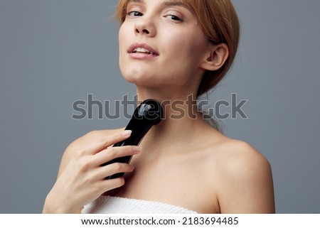 a woman with perfect skin stands on a gray background wrapped in a towel and massages her neck with a black electric anti-aging massager, smiling pleasantly. Close horizontal photo