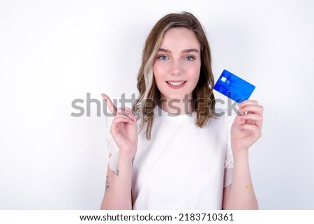 Smiling young caucasian woman wearing white T-shirt over white background showing debit card pointing finger empty space