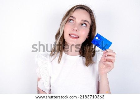 Lovely young caucasian woman wearing white T-shirt over white background showing credit card and looking away at copy space. Pensive