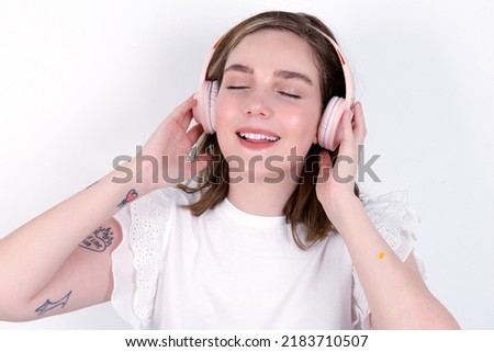 young caucasian woman wearing white T-shirt over white background smiles broadly feels very glad listens favourite music track via wireless headphones closes eyes.
