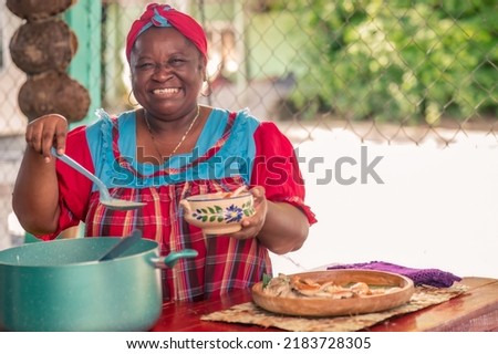 A beautiful African American woman on the patio of her house serving soup. A senior adult woman smiling and looking at the camera as she serves a plate of food.  Royalty-Free Stock Photo #2183728305