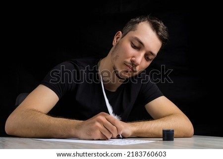 Young handsome and serious man dressed in black quill pen writes on a white sheet of paper on a black background in low key. Royalty-Free Stock Photo #2183760603