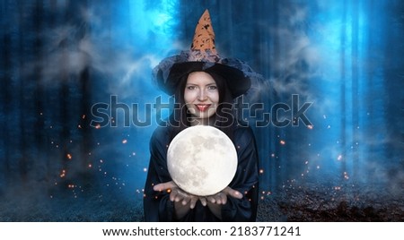 Portrait of smiling witch holding moon on dark blue enchanted foggy forest background.Beautiful young woman in black robe and orange hat with moon in her hands conjuring. Halloween party art design