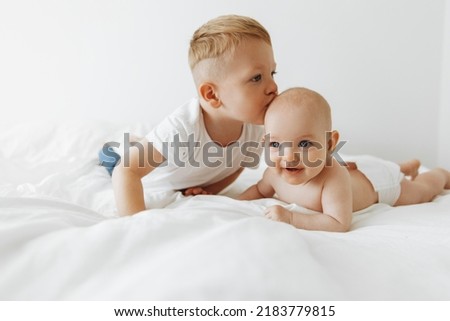 little brother hugs and kisses his newborn sister, a newborn baby. The kid meets a new member of the family. A cute boy and a newborn girl are relaxing in a white bedroom on a bed. Family 