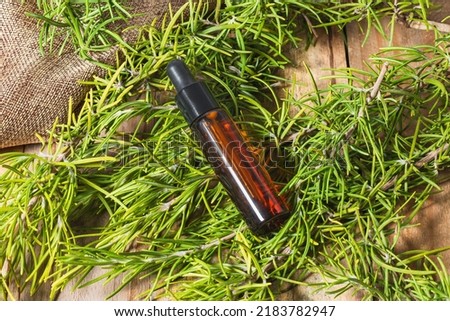 Surrounded by rosemary branches is a brown glass bottle with essential oil capped with a black dropper stopper. Underneath is a sackcloth and wooden planks.