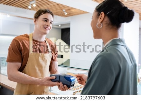 Cheerful seller holding payment terminal near african american client with credit card in sweet shop