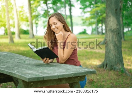 Young woman smiling and reading book in green park outdoors. Novel on nature, concept hobby and lifestyle. Faith, spirituality and religion. Outdoor photo of glad charming girl student.