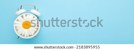 Big beautiful daisy flower on white alarm clock on light blue table background. Pastel color. Time concept. Wide banner. Empty place for text.