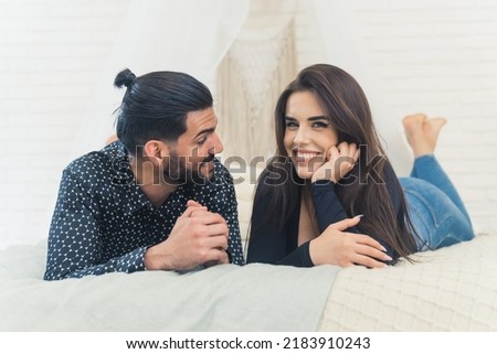 Young white couple relaxing in bedroom together. Boyfriend looking at his girlfriend who's looking at the camera with a wide smile. Indoor shot. High quality photo
