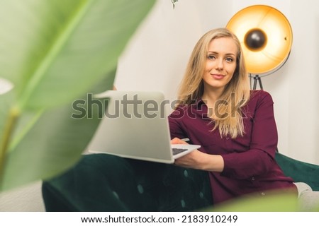 Appealing blonde girl with laptop in her room with a lamp and a plant looking at the camera - room background closeup. High quality photo