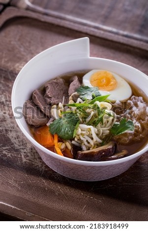 Soup with meat, noodles, boiled egg, parsley, mushrooms, carrots, young shoots and onions in a white plate on a wooden background
