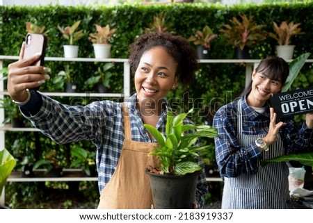Diversity of woman selfie with “welcome we are OPEN” text sign and wearing apron in botanical store. Happy small business owner working at flower shop standing surrounded by plants for customers
