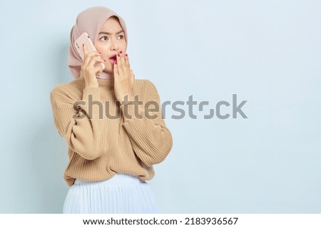 Shocked beautiful Asian muslim woman in brown sweater talking on mobile phone and looking away at copy space isolated over white background. People religious lifestyle concept