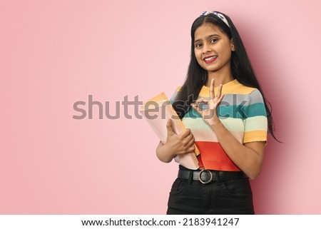 Excited young indian asian girl student standing sideways posing islolated holding textbooks and showing okay sign, looking directly at camera, dark haired female expressing positive emotions.