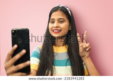 Portrait of pleased nice lovely attractive smiling indian asian girl posing isolated wearing t-shirt doing selfie shot on mobile cell phone  with victory or peace gesture. Mock up copy space.