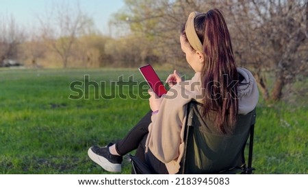 A young girl with dark hair in a beige sweater sits with her back to the camera on a chair and swipes her finger on the smartphone screen, outdoors in the afternoon. 4k