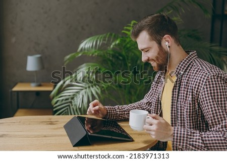 Young happy man he wear shirt earphones work study on tablet pc computer drink tea sit alone at table in coffee shop cafe relax rest in restaurant in free time Freelance mobile office business concept