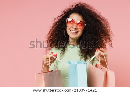 Dreamful fun young curly latin woman 20s wears mint t-shirt sunglasses looking aside holding package bags with purchases after shopping isolated on plain pastel light pink background studio portrait