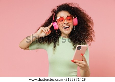 Happy young curly latin woman 20s wears mint t-shirt sunglasses eyes closed listen music in headphones hold in hand use mobile cell phone isolated on plain pastel light pink background studio portrait Royalty-Free Stock Photo #2183971429