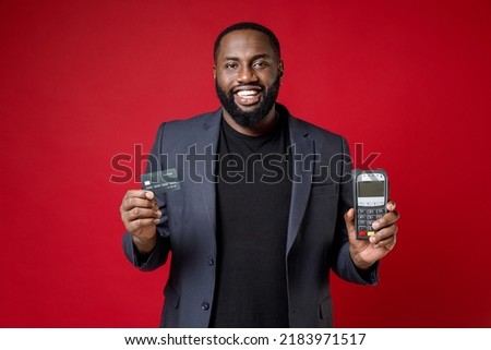 Smiling young african american business man 20s in classic jacket suit hold wireless modern bank payment terminal to process acquire credit card payments isolated on red background studio portrait