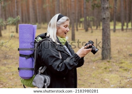 Surprised mother looking at camera screen seeing photos of her family. Elderly woman holding backpack and mat hikking camping trekking in the woods.
