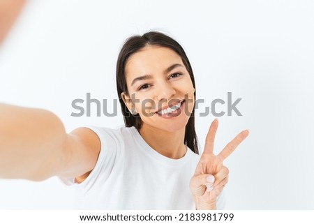 Cheerful brunette young woman making selfie showing two fingers victory gesture and holding camera, white background