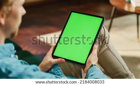 Young Man Holding Tablet Computer with Green Screen Mock Up Display. Male Relaxing at Home, Watching Videos and Reading Social Media Posts on Mobile Device. Close Up Static Over the Shoulder Footage.