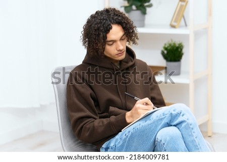 handsome guy writes in a notebook sitting on a chair in the room light background