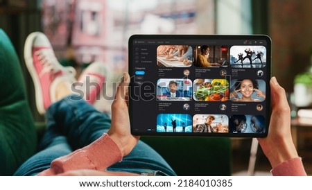 Feminine Hand Holding a Tablet with Video Streaming Service on Display. Female is Relaxing on a Couch at Home, Watching Videos and Reading Social Media Posts on Mobile Device. Close Up POV Photo.
