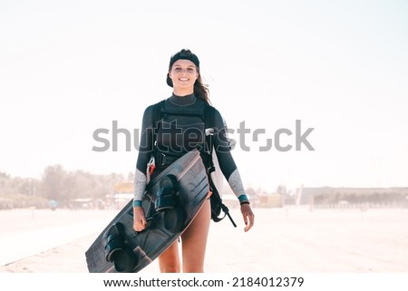 Portrait of beautiful smiling girl posing with a surfboard on the beach and wearing kitesurfing equipment - concept of sporty woman and summer sport activity Royalty-Free Stock Photo #2184012379