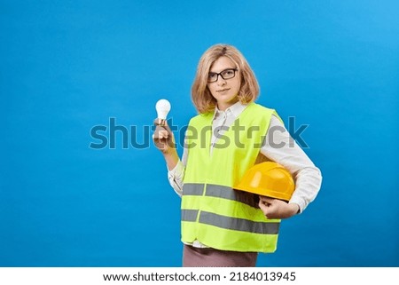Young female engineer wearing work clothes holding a yellow helmet in one hand and holding a light bulb in the other hand while standing in a studio on a blue background