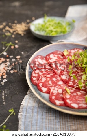 Slices of smoked cervelat salami sausage with spinach microgreen, salt and pepper on black concrete background and beige textile. Side view, close up, selective focus.