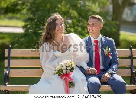 
a young guy in a suit and a girl in a white dress are sitting on a bench and smiling