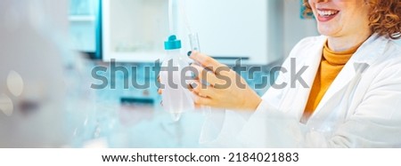 Shot of a female scientist working in a lab. Close-up of serious laboratory worker holding ampoule in front of eyes and examines contents. Scientist in protection gloves eyewear and white coat.