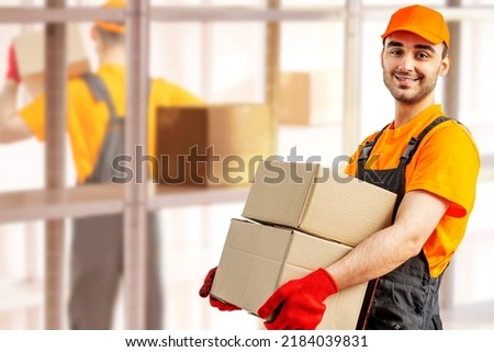Young man holding cardboard package working in warehouse among racks and shelves. Delivery man with box. Staff laborer, orange uniform cap, t-shirt, coveralls service moving delivering orders goods.  Royalty-Free Stock Photo #2184039831