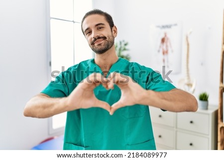 Young physiotherapist working at pain recovery clinic smiling in love doing heart symbol shape with hands. romantic concept. 
