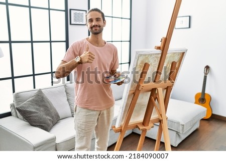 Young hispanic man with beard painting on canvas at home doing happy thumbs up gesture with hand. approving expression looking at the camera showing success. 