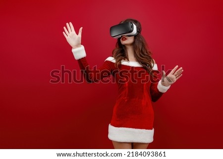 Amazed young Santa girl in Christmas dress looking in headset pointing hands aside isolated on red wall background. Happy New Year 2022 celebration holiday party concept.