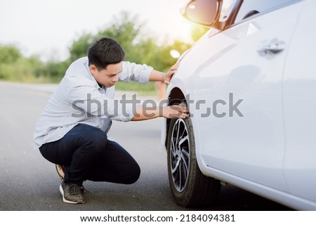 Handsome Asian young man checking wheel flat tire alone on the road. Royalty-Free Stock Photo #2184094381