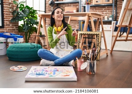 Hispanic woman sitting at art studio painting on canvas smiling and looking at the camera pointing with two hands and fingers to the side. 