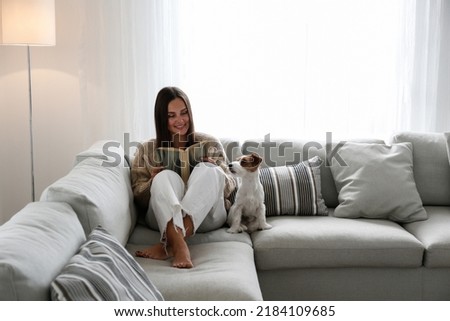 Young beautiful woman reading a book at home with her wire haired jack russell terrier pup. Brunette female and rough coated dog sitting on textile couch. White wall background, copy space, close up. Royalty-Free Stock Photo #2184109685
