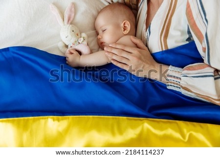 Close-up portrait of a mother kissing her newborn baby sleeping on the bed at home covered with the Blue and yellow flag of Ukraine. The baby sleeps with the safety and protection of the mother