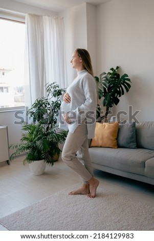 Full length view of the beautiful pregnant woman standing near window at home and looking at the distance. Waiting for the baby concept. Stock photo