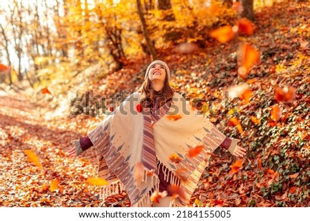 Cheerful young woman having fun spending sunny autumn day in nature, looking up with arms spread, watching the colorful leaves fall on the forest path