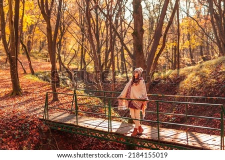 Woman walking down the bridge over a stream on the forest path covered with colorful fallen leaves, enjoying spending sunny autumn day outdoors in nature Royalty-Free Stock Photo #2184155019