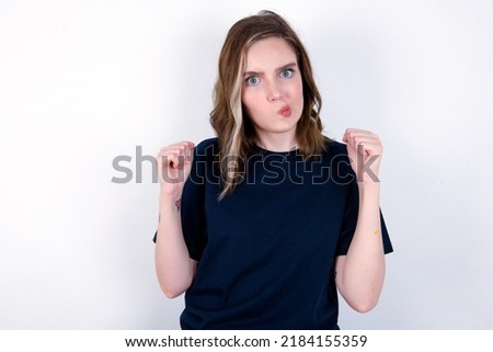 Irritated young caucasian woman wearing black T-shirt over white background blows cheeks with anger and raises clenched fists expresses rage and aggressive emotions. Furious model Royalty-Free Stock Photo #2184155359